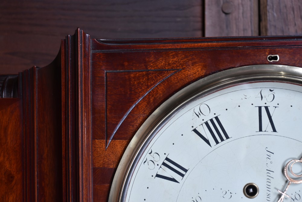 An antique Scottish flame mahogany longcase clock featuring a carved swan-neck pediment with brass finials, a rounded dial, and a veneered trunk door. View XIV
