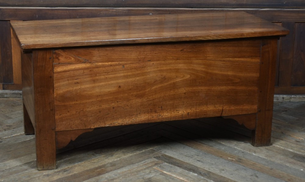 An antique French cherrywood blanket box with a hinged rectangular lid, natural wood grain, and shaped corner apron.
