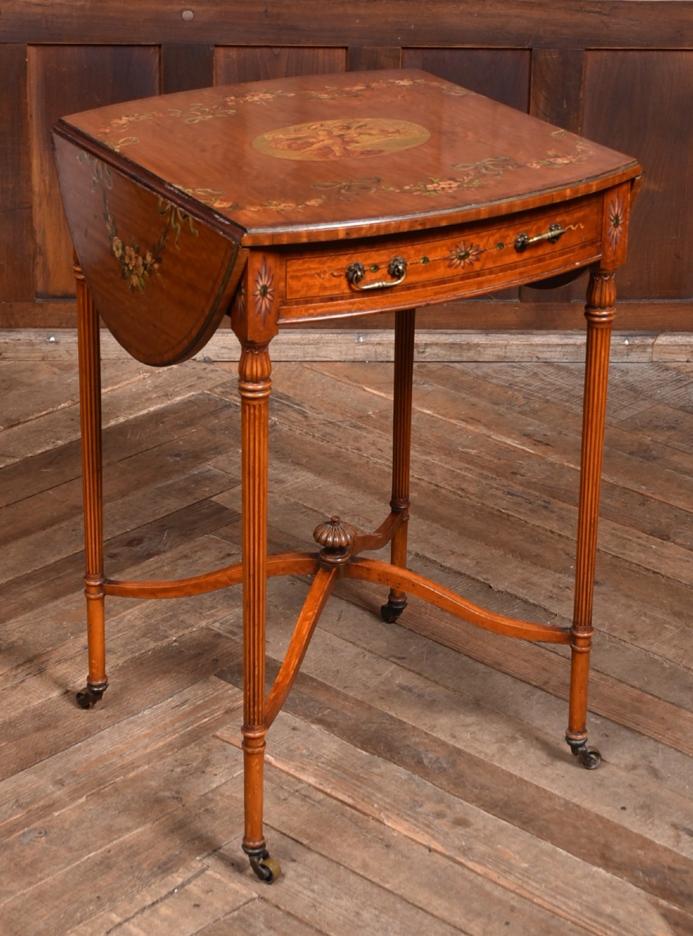 An Edwardian Neoclassical drop-leaf table featuring hand-painted floral swags, a central figural medallion, fluted turned legs, and an X-stretcher on castors. View II