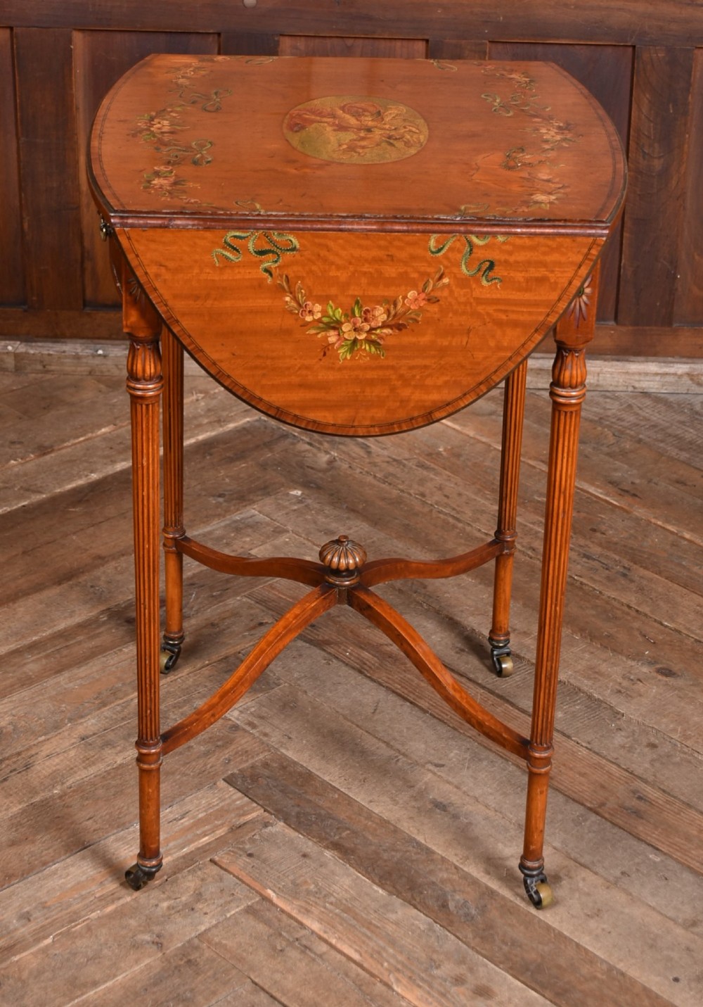 An Edwardian Neoclassical drop-leaf table featuring hand-painted floral swags, a central figural medallion, fluted turned legs, and an X-stretcher on castors. View V