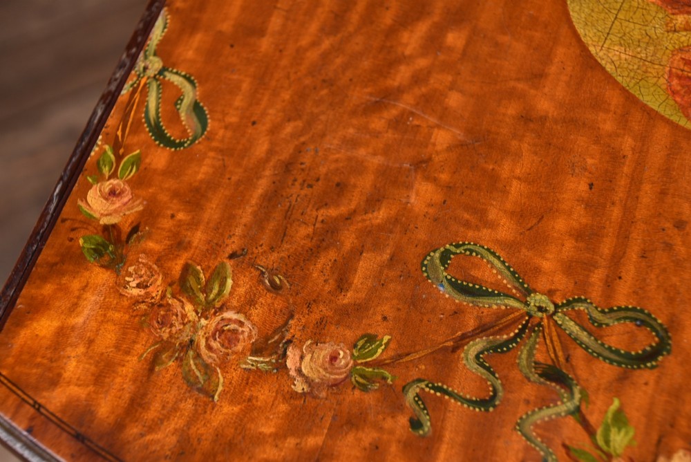 An Edwardian Neoclassical drop-leaf table featuring hand-painted floral swags, a central figural medallion, fluted turned legs, and an X-stretcher on castors. View X