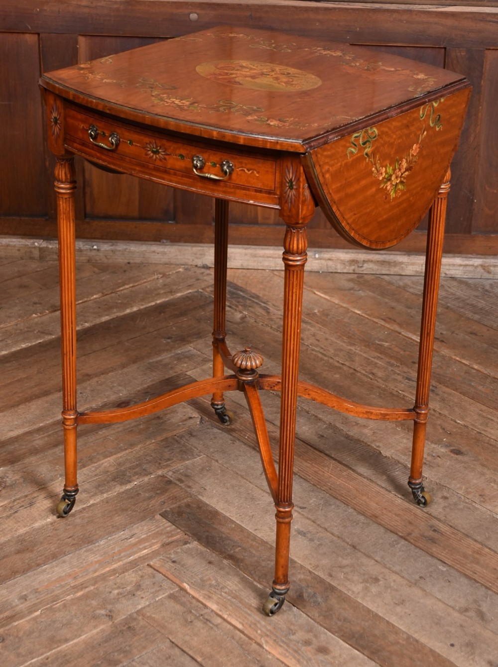 An Edwardian Neoclassical drop-leaf table featuring hand-painted floral swags, a central figural medallion, fluted turned legs, and an X-stretcher on castors. View XV