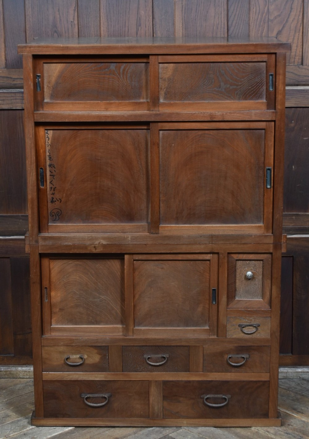 A tall wooden Japanese Mizuya tansu cabinet featuring multiple sliding door compartments, small drawers with iron drop handles, and faint calligraphic inscriptions on an upper panel.