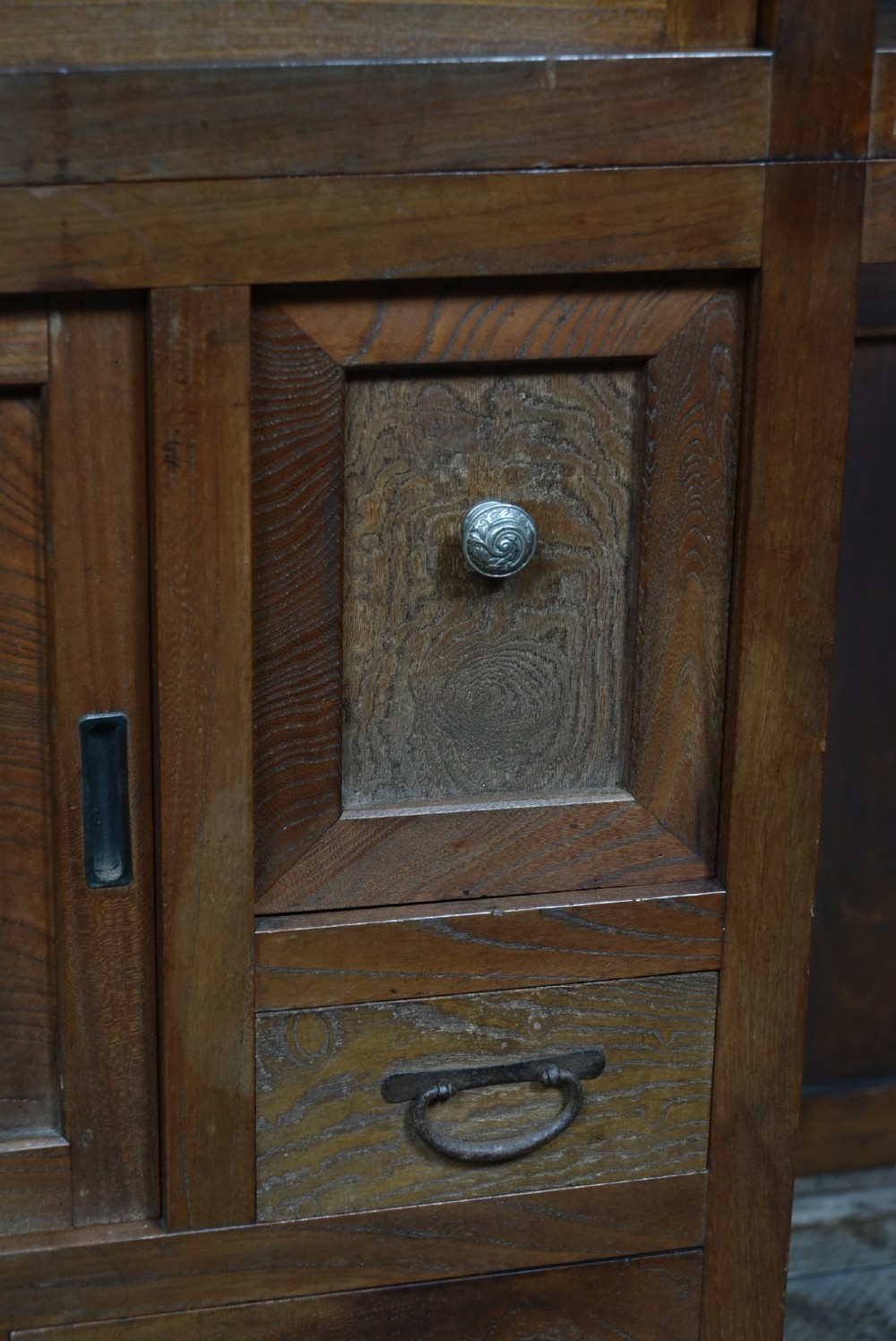 A tall wooden Japanese Mizuya tansu cabinet featuring multiple sliding door compartments, small drawers with iron drop handles, and faint calligraphic inscriptions on an upper panel. View III