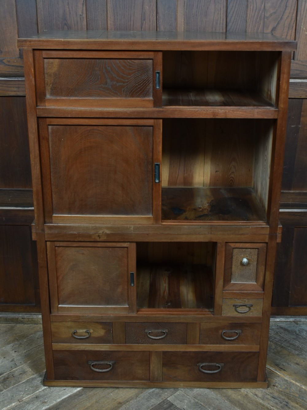 A tall wooden Japanese Mizuya tansu cabinet featuring multiple sliding door compartments, small drawers with iron drop handles, and faint calligraphic inscriptions on an upper panel. View V