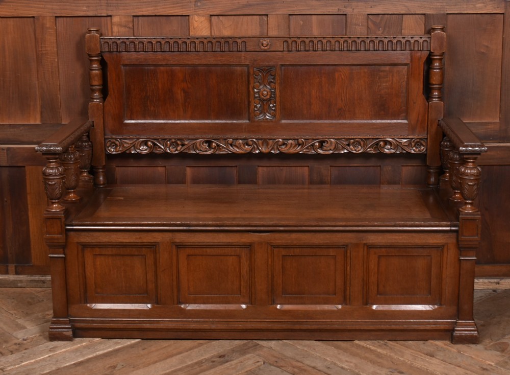 A dark carved oak hall bench featuring a panelled backrest, turned arm supports, and a hinged box storage seat.