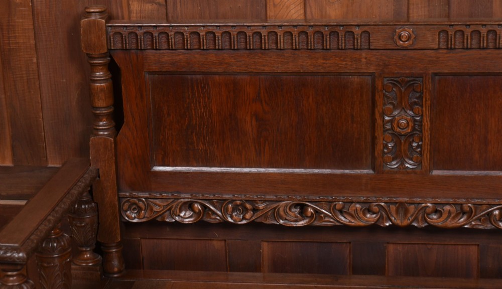 A dark carved oak hall bench featuring a panelled backrest, turned arm supports, and a hinged box storage seat. View III