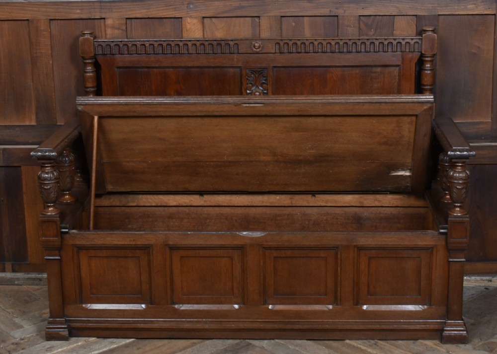 A dark carved oak hall bench featuring a panelled backrest, turned arm supports, and a hinged box storage seat. View IV