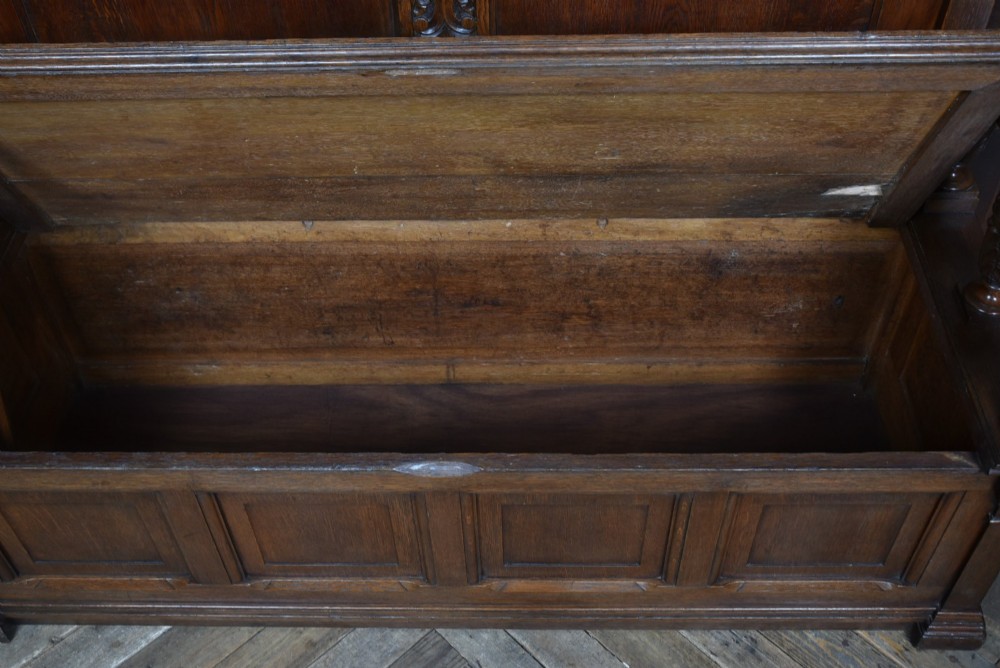 A dark carved oak hall bench featuring a panelled backrest, turned arm supports, and a hinged box storage seat. View V