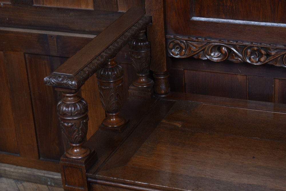 A dark carved oak hall bench featuring a panelled backrest, turned arm supports, and a hinged box storage seat. View VI
