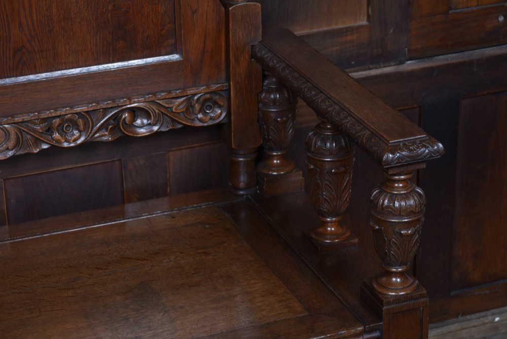 A dark carved oak hall bench featuring a panelled backrest, turned arm supports, and a hinged box storage seat. View VII