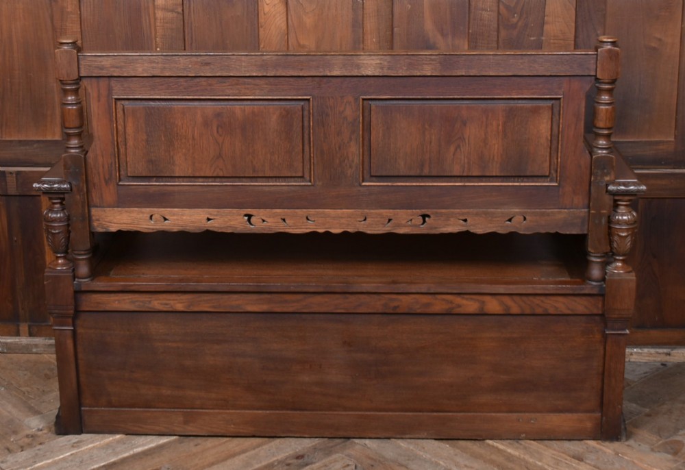 A dark carved oak hall bench featuring a panelled backrest, turned arm supports, and a hinged box storage seat. View XII
