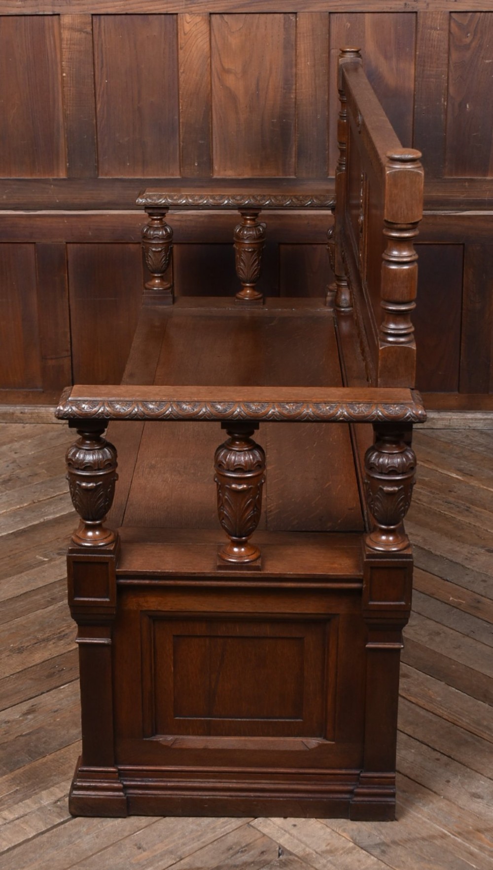 A dark carved oak hall bench featuring a panelled backrest, turned arm supports, and a hinged box storage seat. View XIII