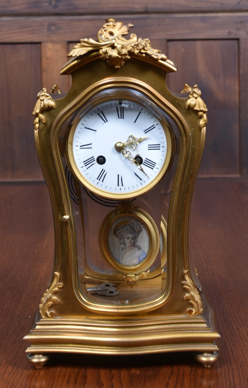 A 19th-century French gilt bronze mantel clock featuring curved glass panels, a white enamel dial with Roman numerals, and an oval portrait miniature pendulum signed Rodier.