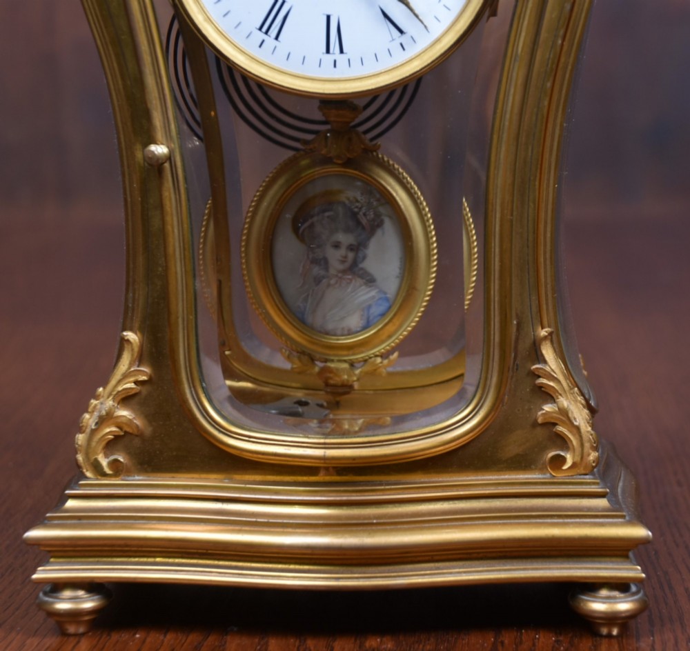 A 19th-century French gilt bronze mantel clock featuring curved glass panels, a white enamel dial with Roman numerals, and an oval portrait miniature pendulum signed Rodier. View II