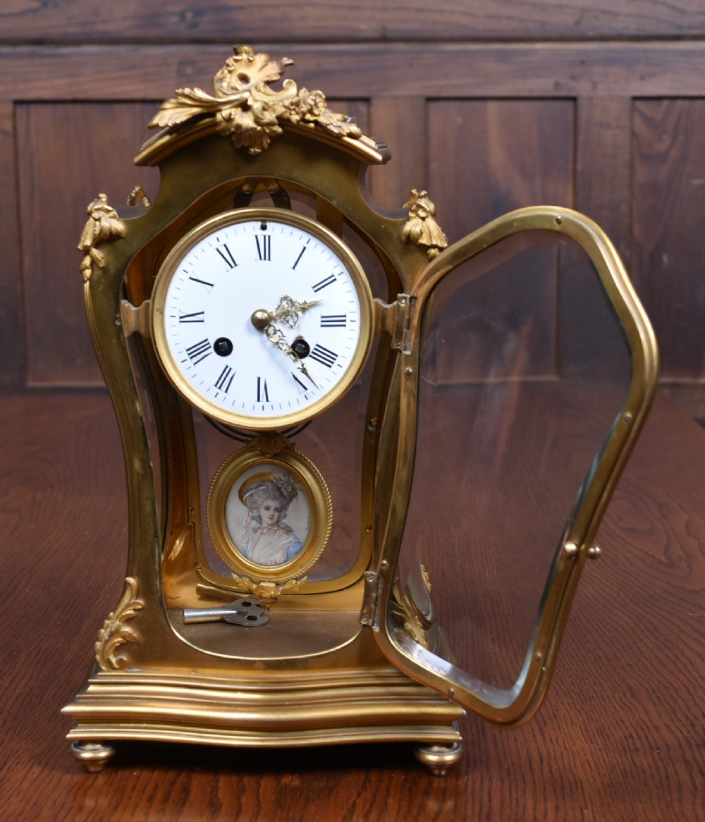 A 19th-century French gilt bronze mantel clock featuring curved glass panels, a white enamel dial with Roman numerals, and an oval portrait miniature pendulum signed Rodier. View IV