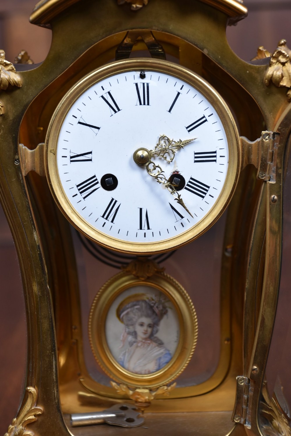 A 19th-century French gilt bronze mantel clock featuring curved glass panels, a white enamel dial with Roman numerals, and an oval portrait miniature pendulum signed Rodier. View V