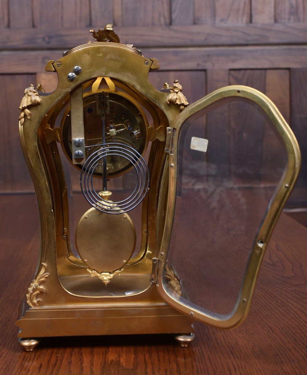 A 19th-century French gilt bronze mantel clock featuring curved glass panels, a white enamel dial with Roman numerals, and an oval portrait miniature pendulum signed Rodier. View VIII