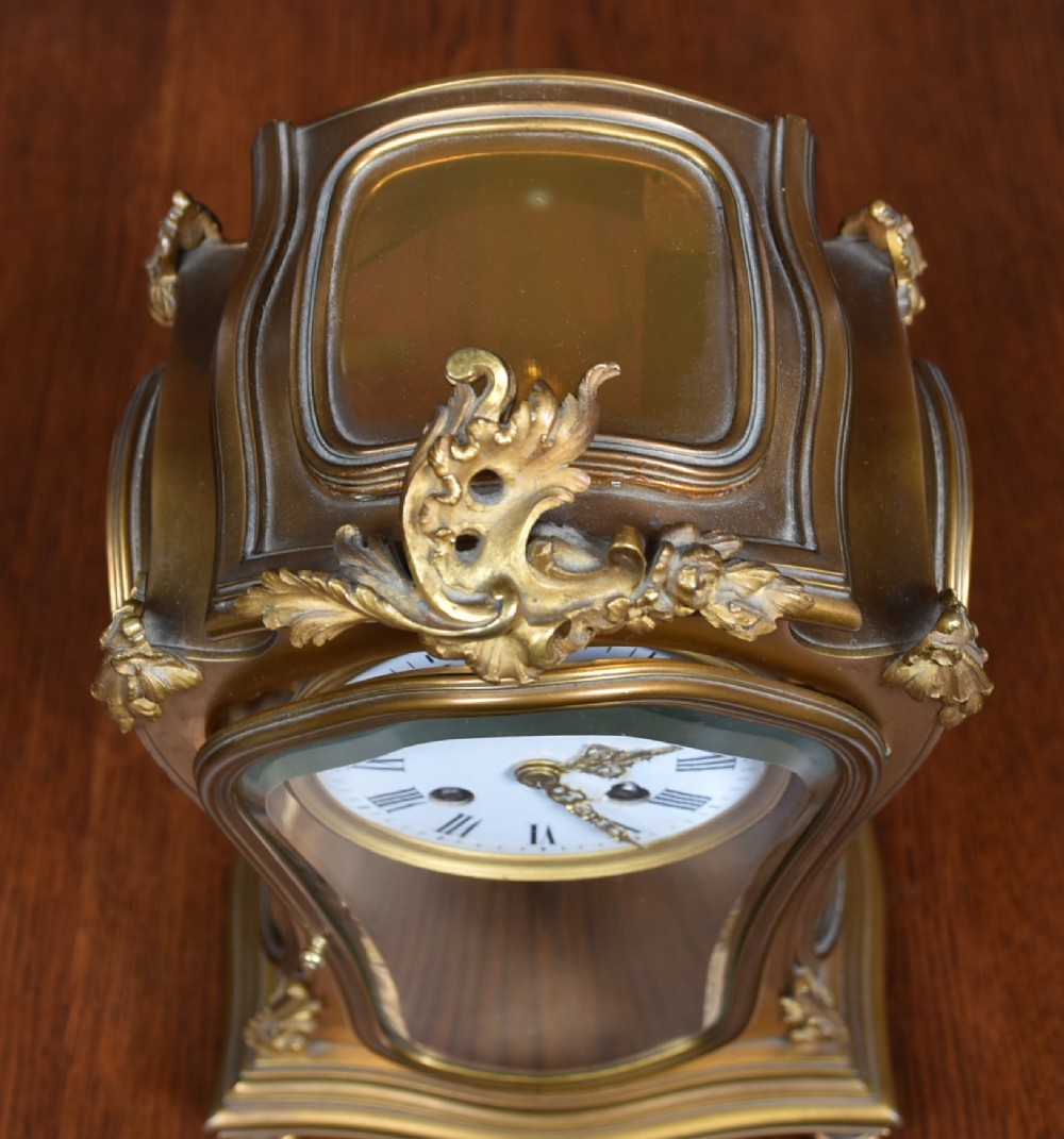 A 19th-century French gilt bronze mantel clock featuring curved glass panels, a white enamel dial with Roman numerals, and an oval portrait miniature pendulum signed Rodier. View XI