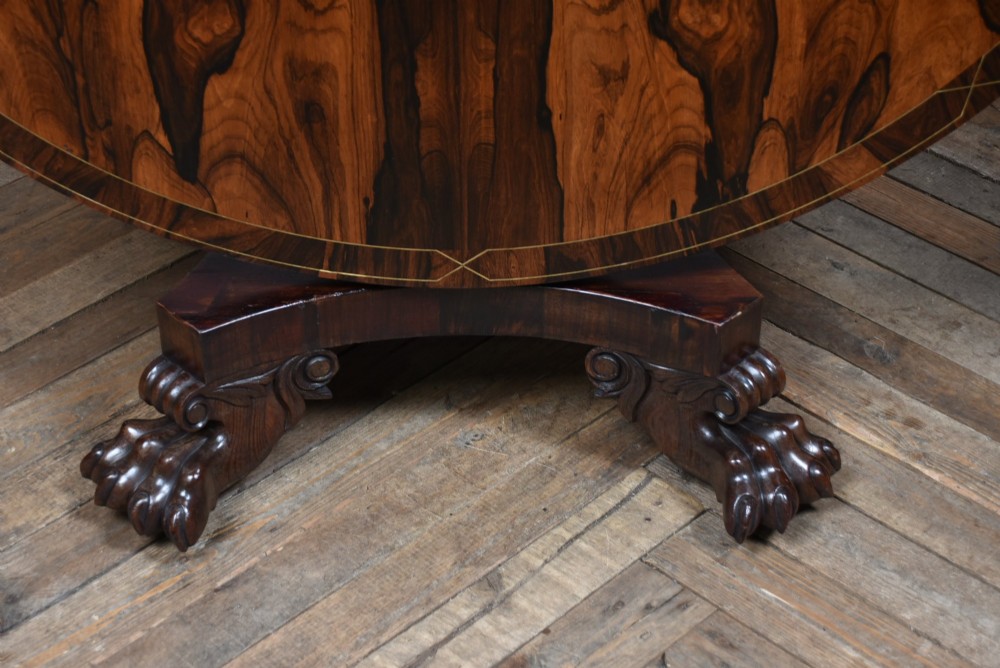 A circular Victorian rosewood tilt-top table featuring highly figured timber grain, a deep apron, a carved central pedestal column, and a quadform base on carved claw feet. View VI