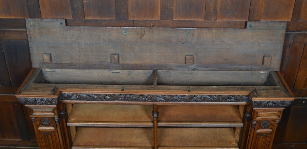 A wide 19th-century Victorian Gothic Revival open oak bookcase featuring detailed floral carvings, moulded pilasters, and twin bays of adjustable wooden shelves. View XI