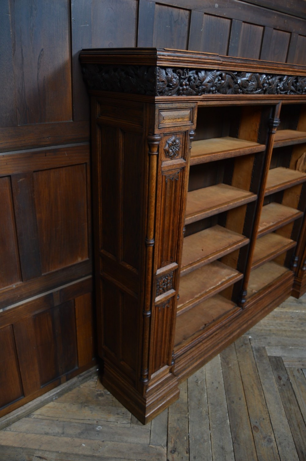 A wide 19th-century Victorian Gothic Revival open oak bookcase featuring detailed floral carvings, moulded pilasters, and twin bays of adjustable wooden shelves. View XIV