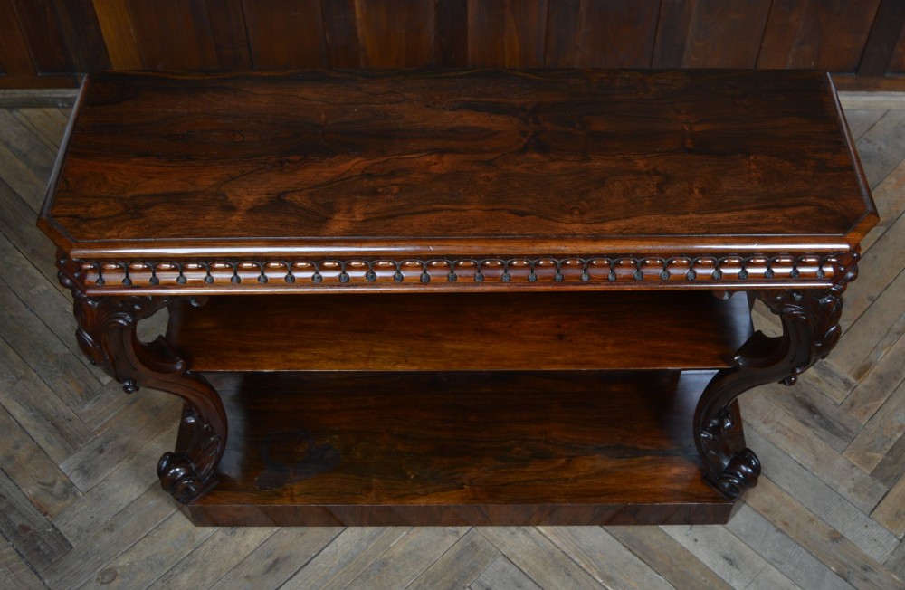A dark Victorian rosewood console table featuring carved S-scroll supports, an open central shelf, a beaded upper frieze, and a solid plinth base. View VI