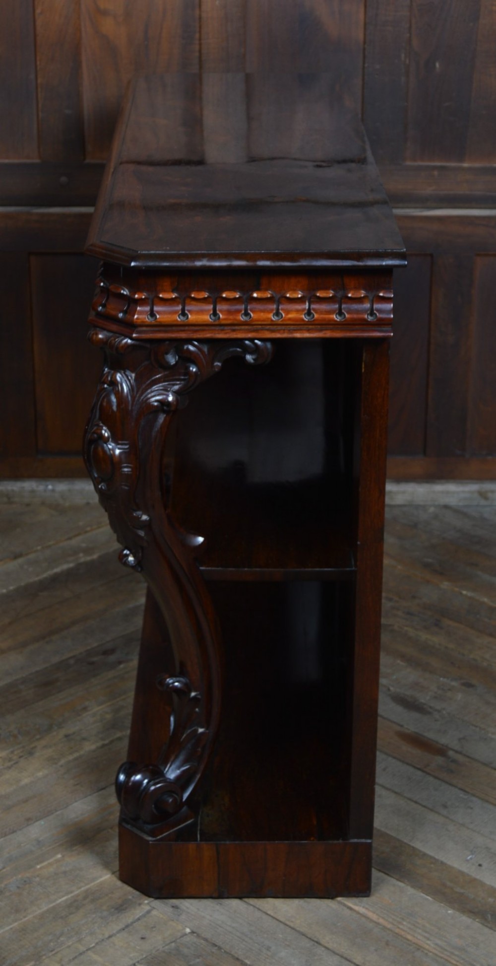 A dark Victorian rosewood console table featuring carved S-scroll supports, an open central shelf, a beaded upper frieze, and a solid plinth base. View X