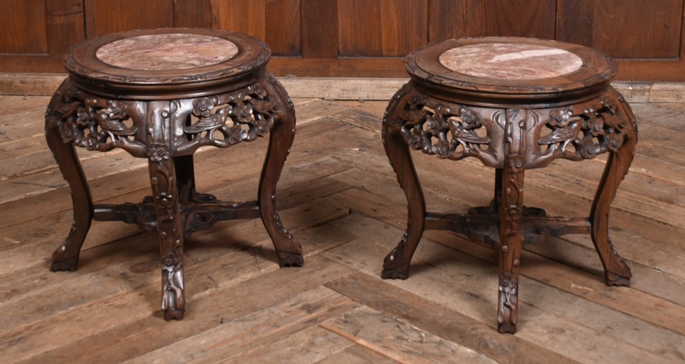 A pair of carved dark hardwood round stands featuring inset pink marble tops and pierced floral aprons supported by curved legs.