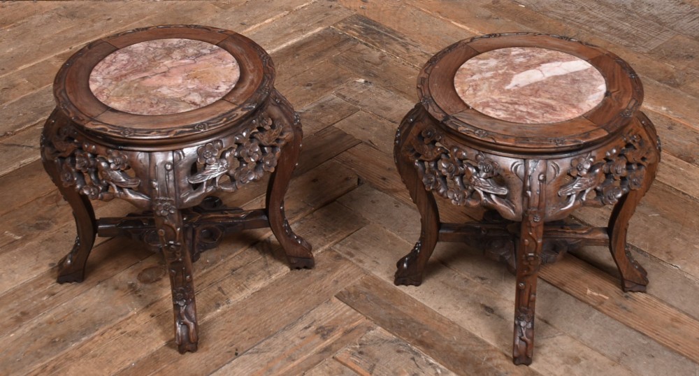 A pair of carved dark hardwood round stands featuring inset pink marble tops and pierced floral aprons supported by curved legs. View II