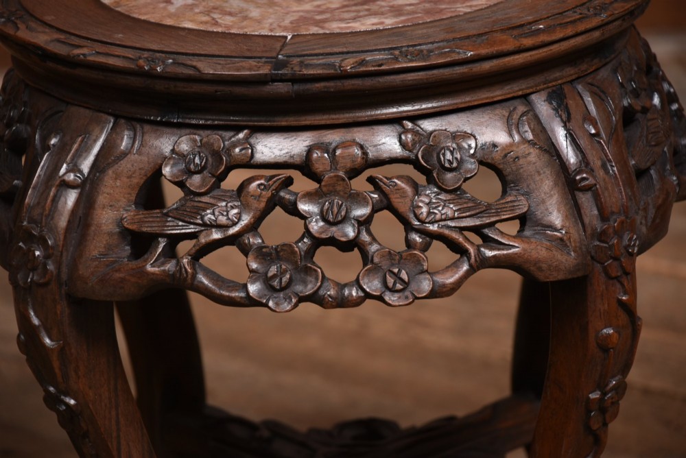 A pair of carved dark hardwood round stands featuring inset pink marble tops and pierced floral aprons supported by curved legs. View X