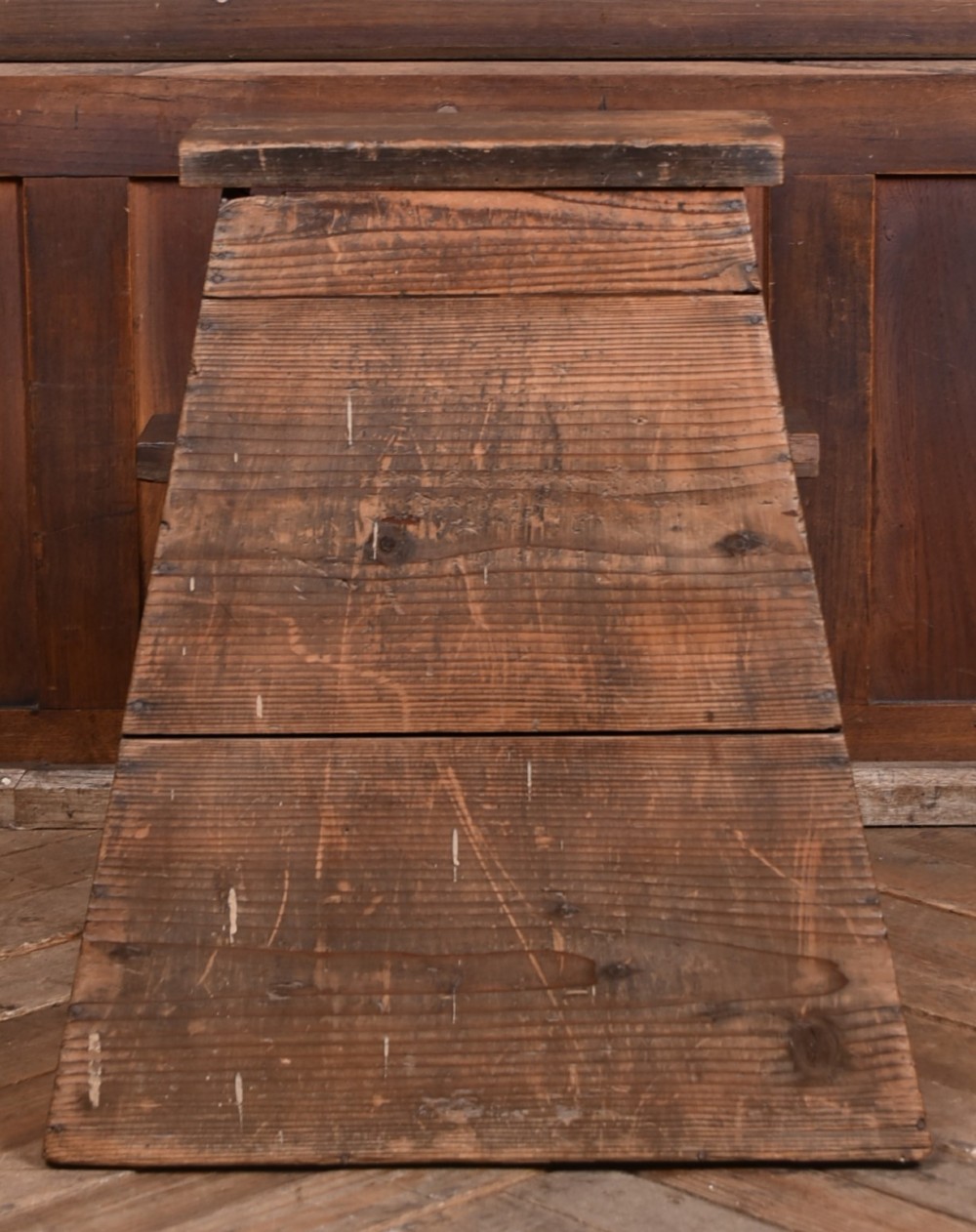 A weathered brown wooden step stool with a tapered rectangular base and an upper platform seat displaying distinct grain and patination. View V