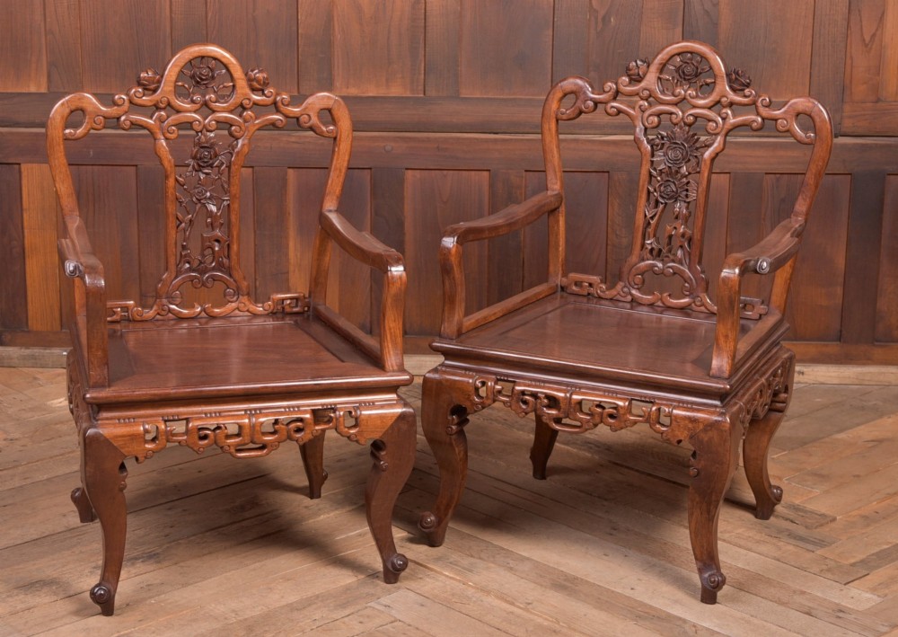 A pair of dark warm brown Chinese hardwood armchairs featuring intricately carved floral back splats, openwork scroll aprons, and sculpted legs.