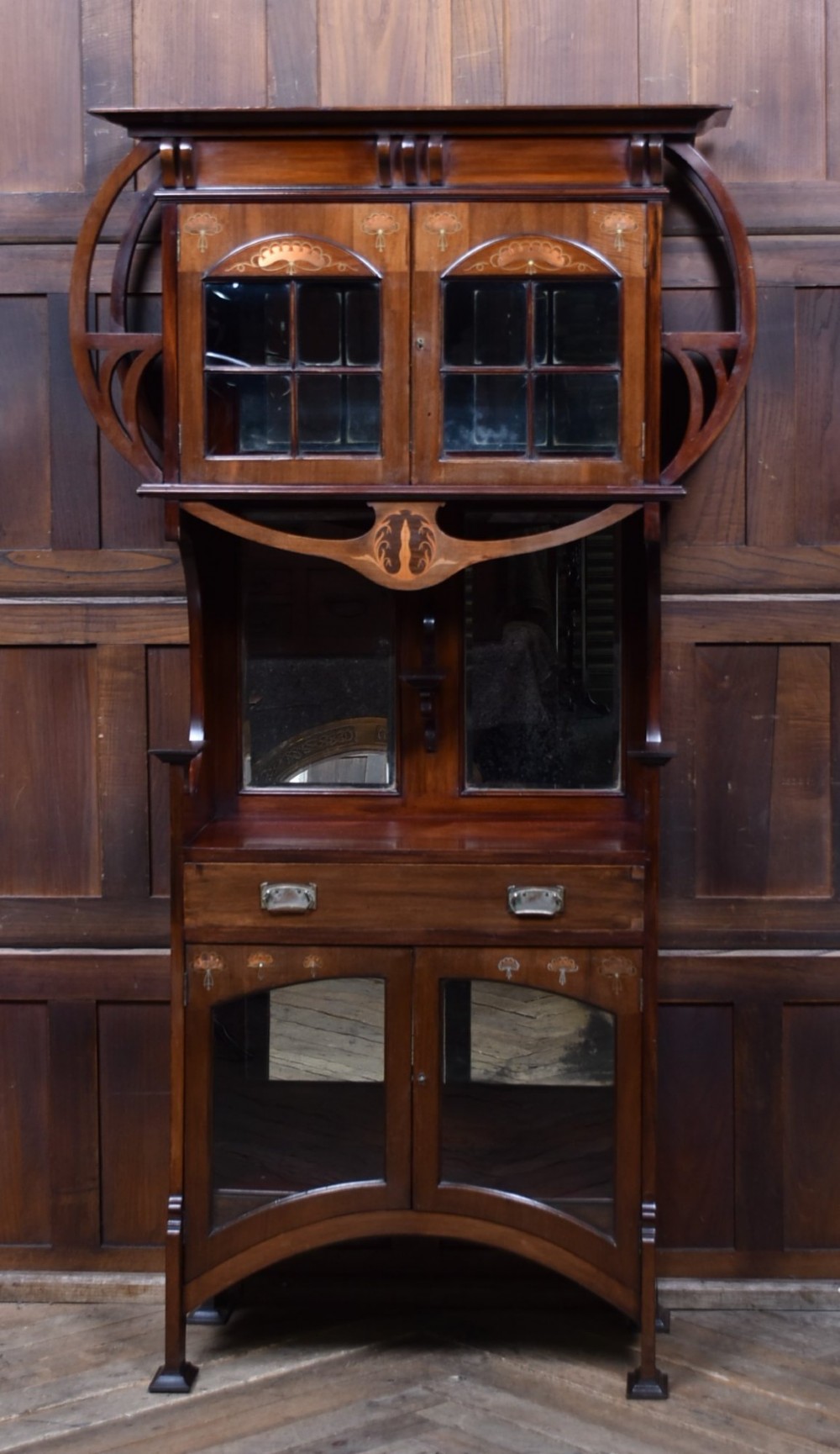 An Art Nouveau dark mahogany display cabinet featuring botanical inlay motifs, glazed upper doors, a central mirrored recess, a single drawer, and curved side brackets.