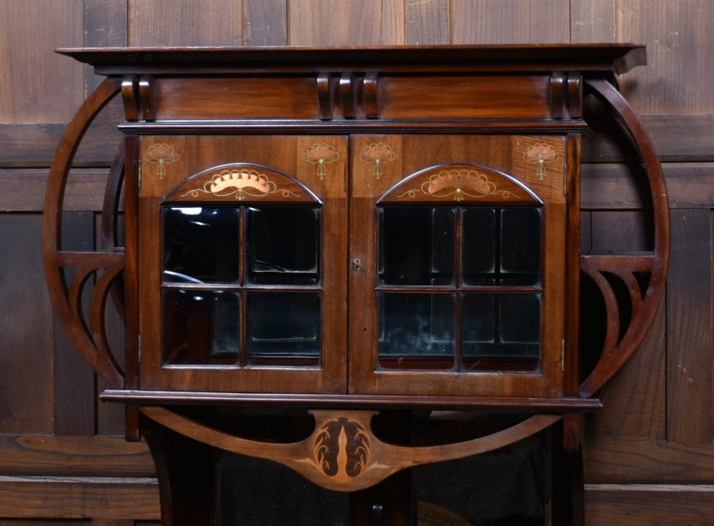An Art Nouveau dark mahogany display cabinet featuring botanical inlay motifs, glazed upper doors, a central mirrored recess, a single drawer, and curved side brackets. View II