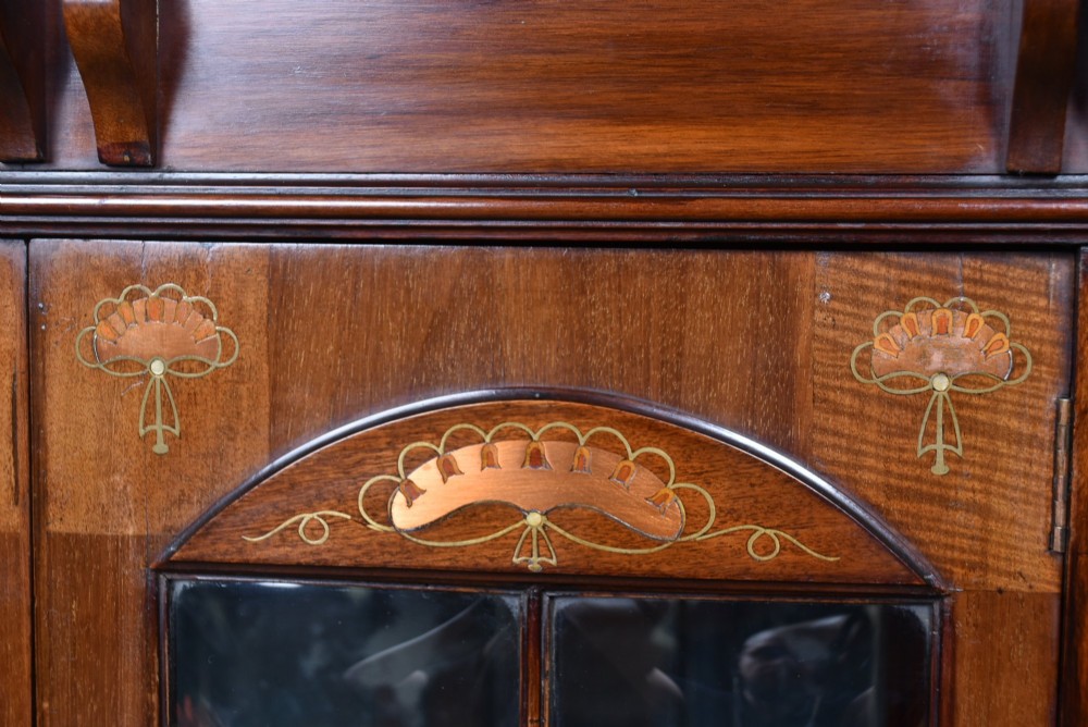 An Art Nouveau dark mahogany display cabinet featuring botanical inlay motifs, glazed upper doors, a central mirrored recess, a single drawer, and curved side brackets. View III