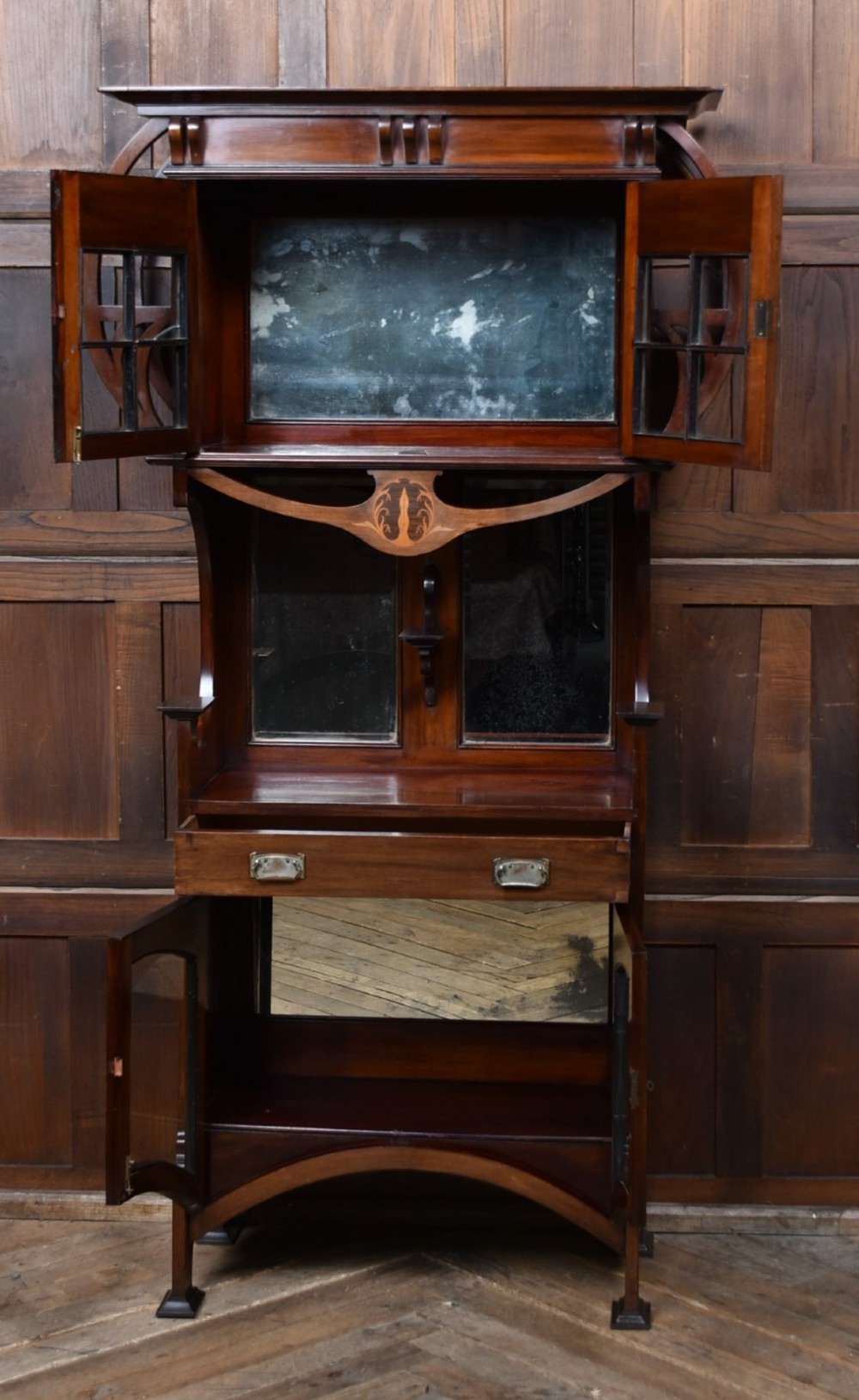 An Art Nouveau dark mahogany display cabinet featuring botanical inlay motifs, glazed upper doors, a central mirrored recess, a single drawer, and curved side brackets. View IV
