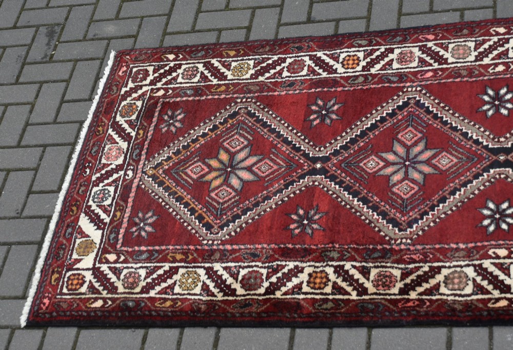 A long handwoven wool runner rug featuring five central diamond medallions on a dark red ground with geometric borders. View IV