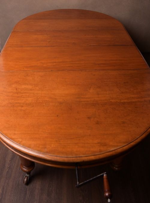 A Victorian mahogany extendable dining table featuring turned legs on brass castors, rounded tabletop corners, and an exposed metal winding crank handle. View VII