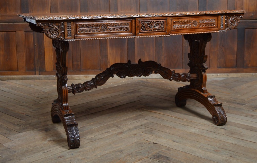 A Victorian carved padauk wood library table featuring three frieze drawers and an intricate carved trestle base with an arched central stretcher.