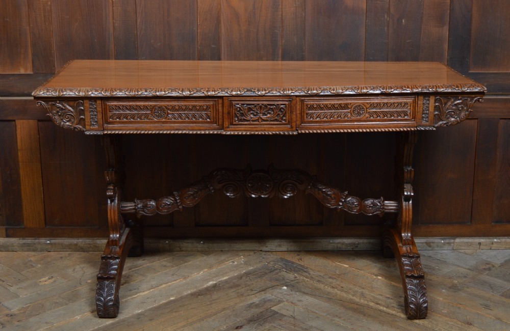 A Victorian carved padauk wood library table featuring three frieze drawers and an intricate carved trestle base with an arched central stretcher. View II
