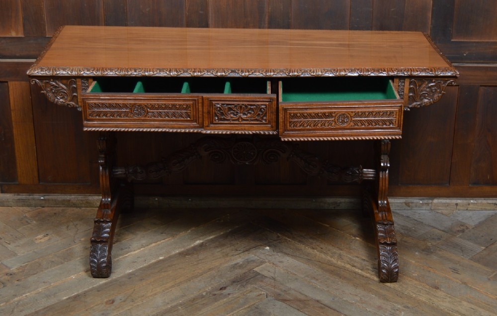 A Victorian carved padauk wood library table featuring three frieze drawers and an intricate carved trestle base with an arched central stretcher. View III