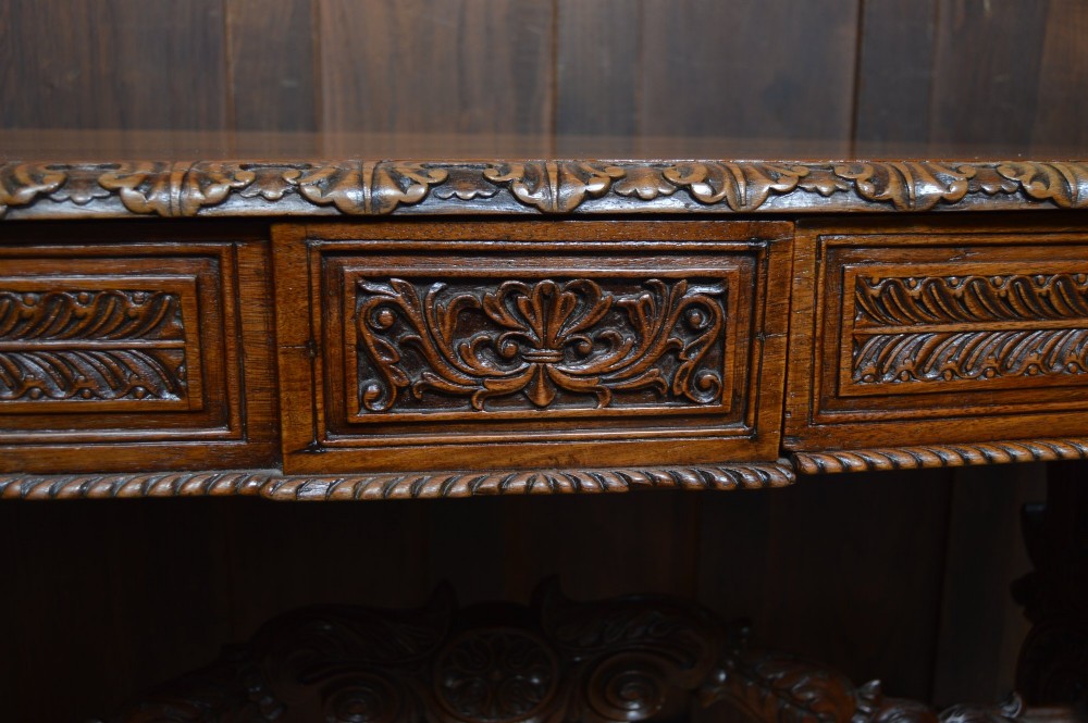 A Victorian carved padauk wood library table featuring three frieze drawers and an intricate carved trestle base with an arched central stretcher. View IV
