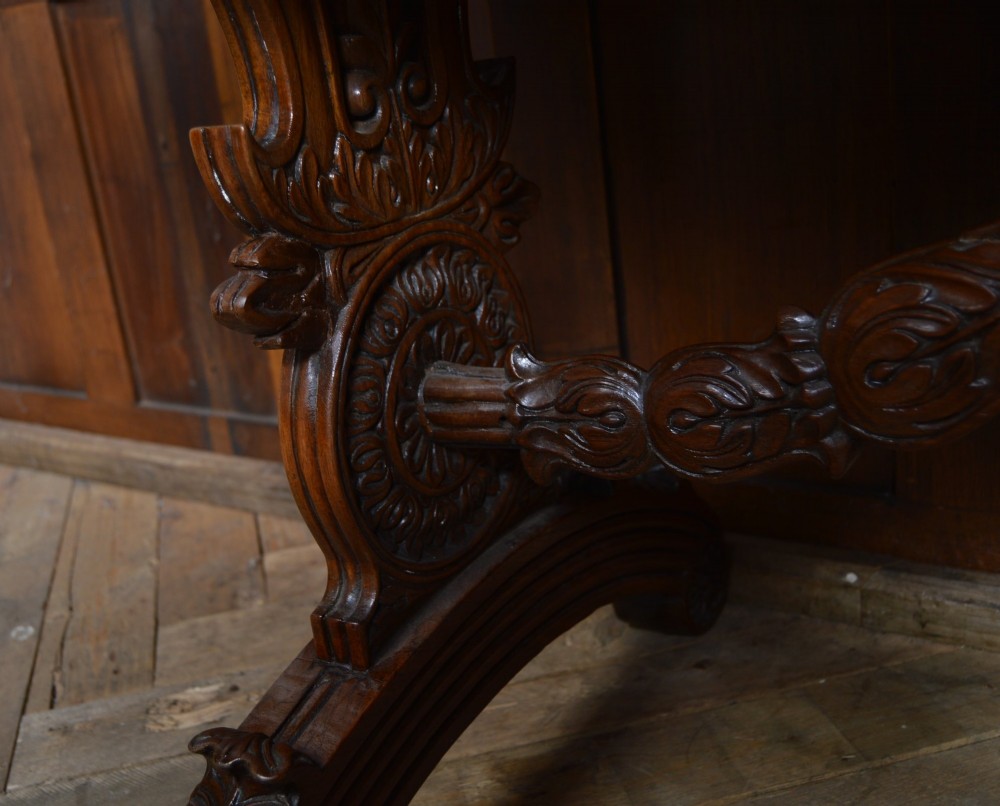 A Victorian carved padauk wood library table featuring three frieze drawers and an intricate carved trestle base with an arched central stretcher. View VI