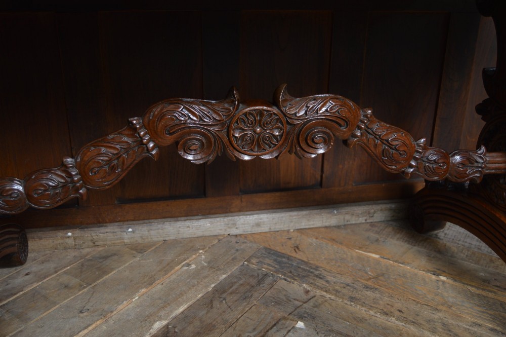 A Victorian carved padauk wood library table featuring three frieze drawers and an intricate carved trestle base with an arched central stretcher. View VII
