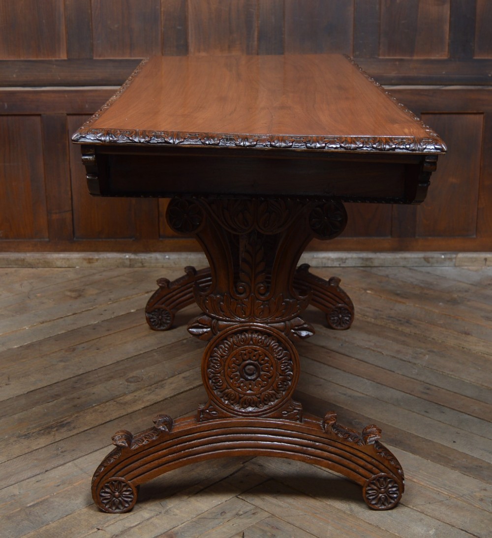 A Victorian carved padauk wood library table featuring three frieze drawers and an intricate carved trestle base with an arched central stretcher. View VIII