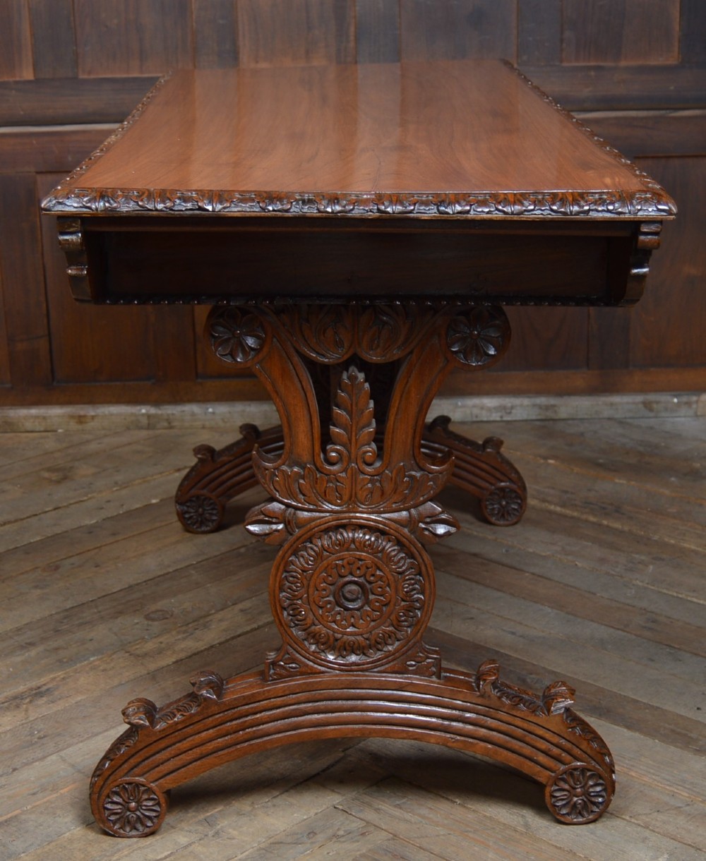 A Victorian carved padauk wood library table featuring three frieze drawers and an intricate carved trestle base with an arched central stretcher. View IX