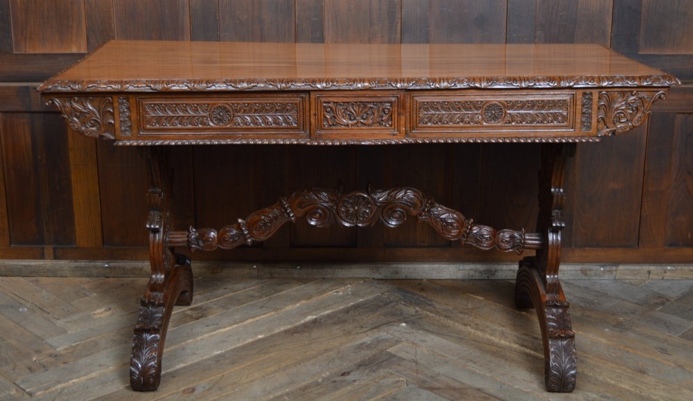 A Victorian carved padauk wood library table featuring three frieze drawers and an intricate carved trestle base with an arched central stretcher. View X