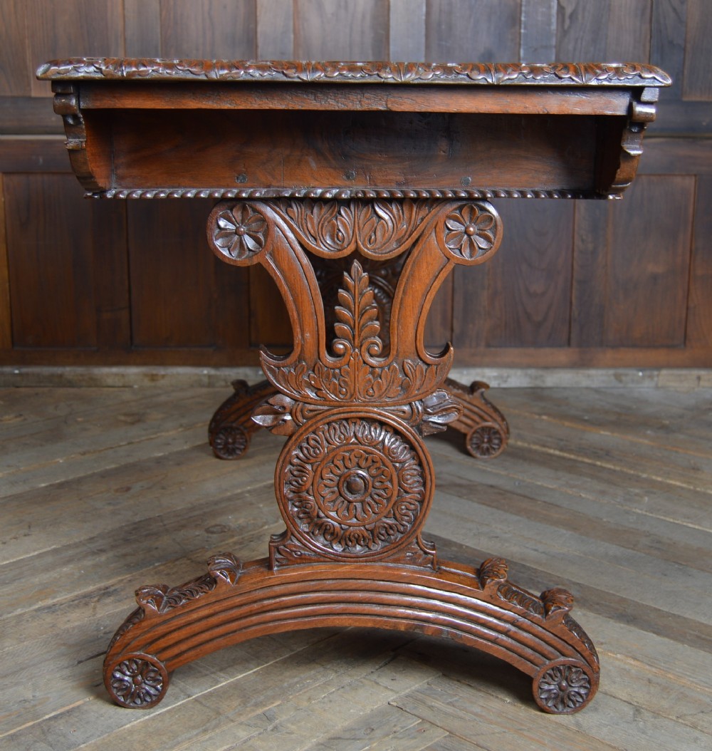 A Victorian carved padauk wood library table featuring three frieze drawers and an intricate carved trestle base with an arched central stretcher. View XI