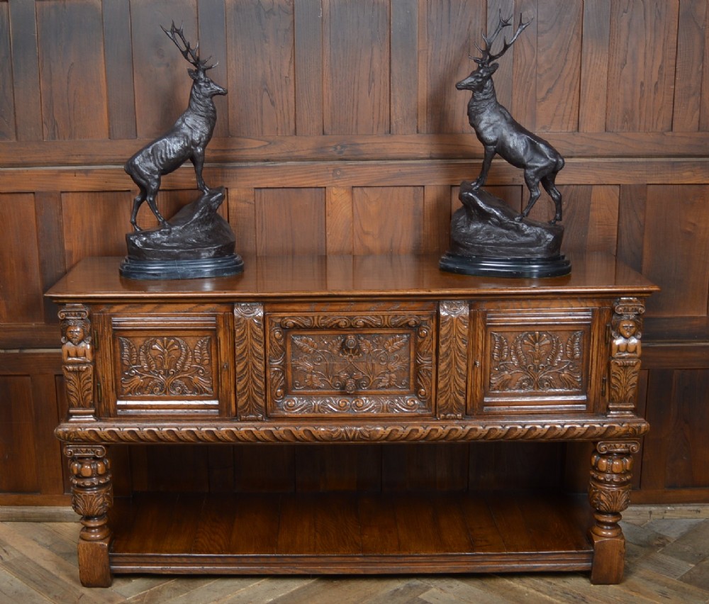A carved dark oak Edwardian pot board dresser base featuring detailed relief carving on cupboard doors and a wide lower open shelf.